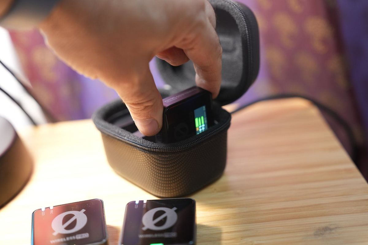A hand places a small electronic device with a screen displaying green bars into a black zippered case on a wooden surface.