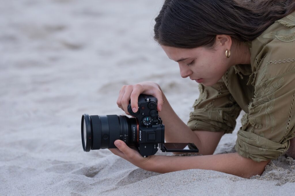 A woman kneelling on the sand, holding the Sony a7 V while recording using the viewfinder. 